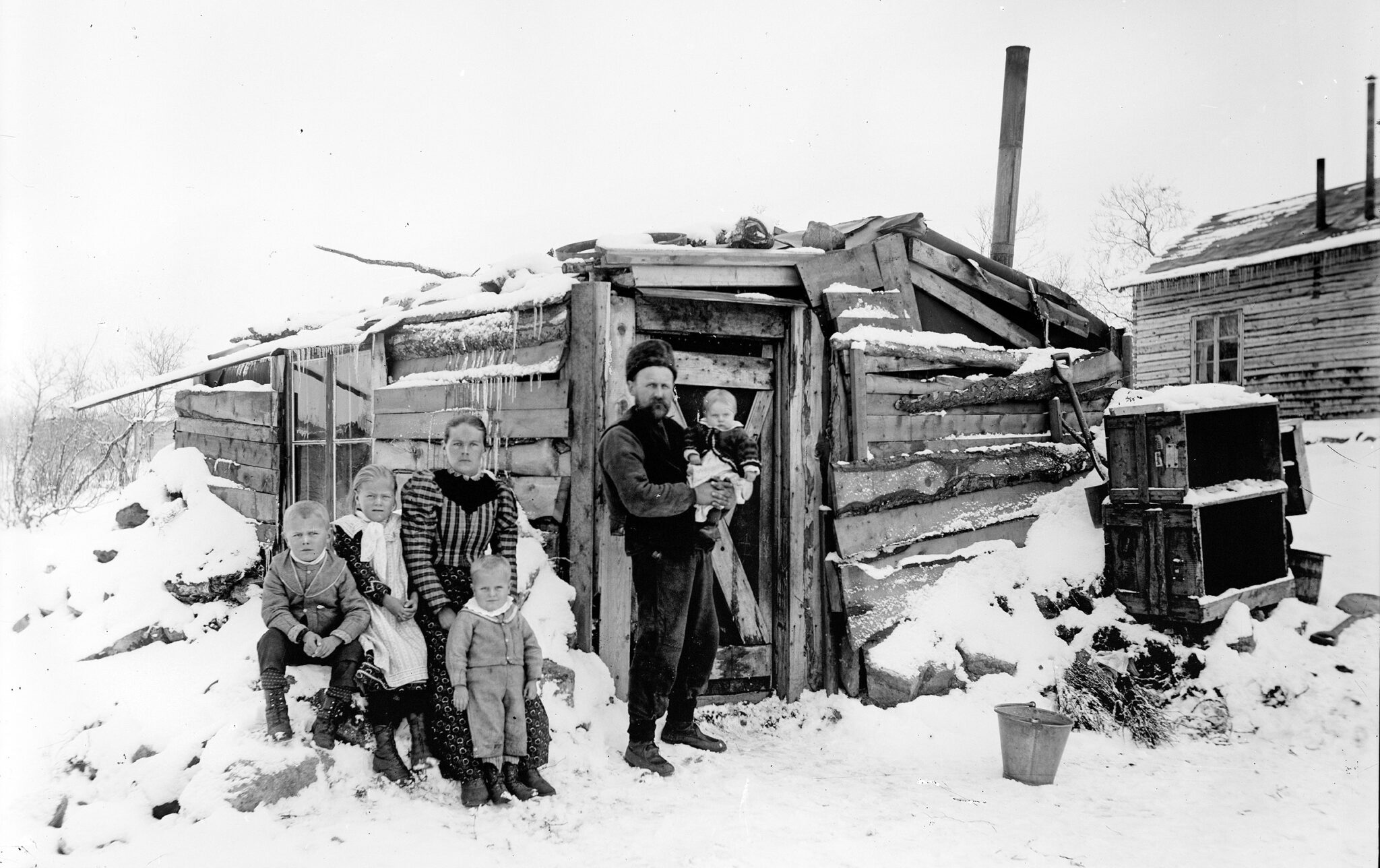 Ingeborg och Anders Söderberg Borg Mesch, tillsammans med sina fyra barn står framför sitt enkla hus byggt av restplank. I bakgrunden syns ett timrat boningshus. Landskapet är snötäckt.