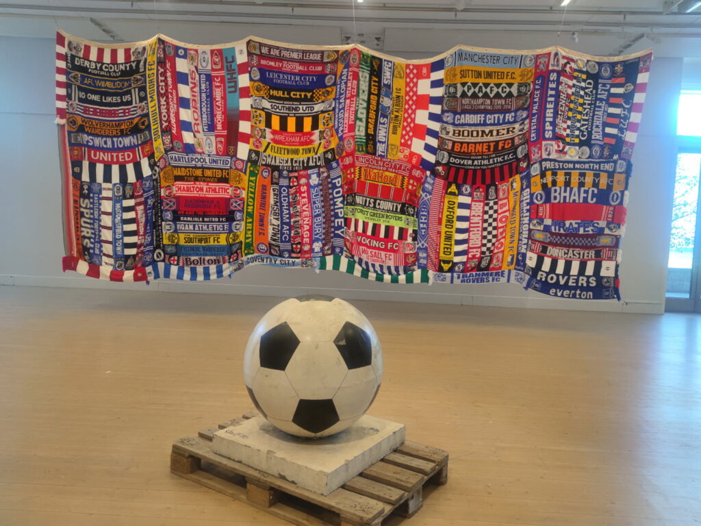 Utställning med fotbollstema, olika lags halsdukar är upphängda bakom en skulptur av en fotboll.
